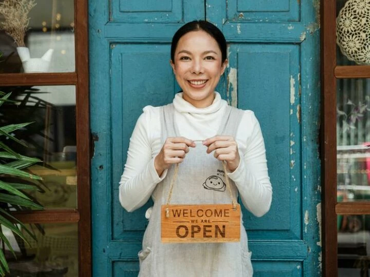 Cheerful ethnic female cafeteria owner in apron demonstrating cardboard signboard while standing near blue shabby door and windows after starting own business and looking at camera