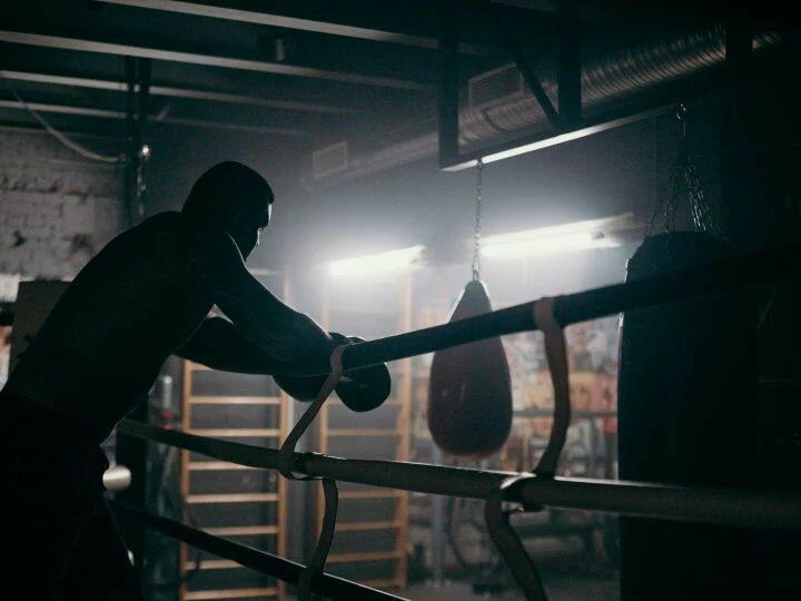 Moody silhouette of a boxer leaning in a dimly lit gym with punching bag and boxing ring.