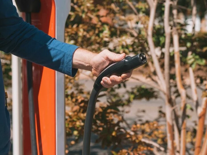 Hand holding an electric vehicle charger at a station outdoors with autumn trees.